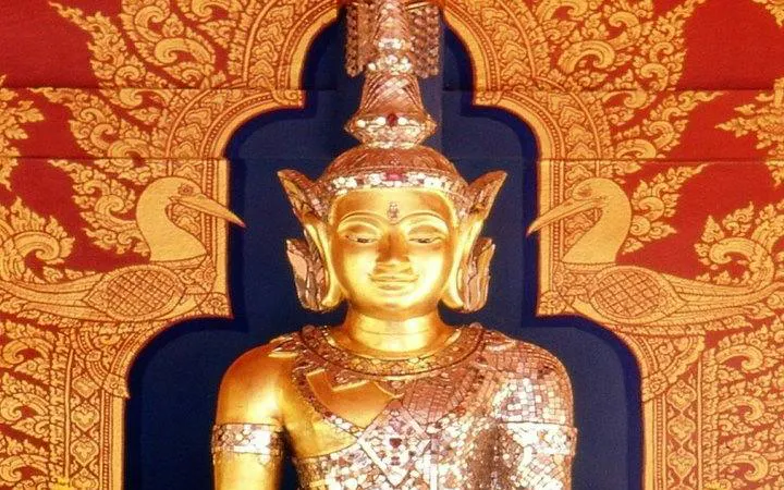 Golden Buddha statue at Wat Phra Kaew, Chiang Rai, with intricate red and gold backdrop.