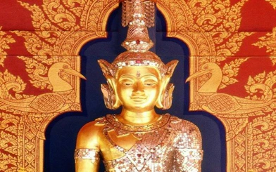 Golden Buddha statue at Wat Phra Kaew, Chiang Rai, with intricate red and gold backdrop.