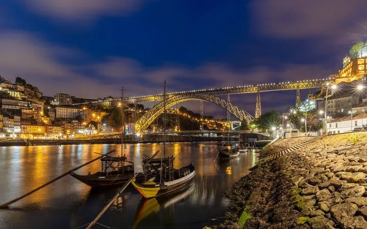 Porto's Dom Luís I Bridge lit up at night over the Douro River with traditional boats.
