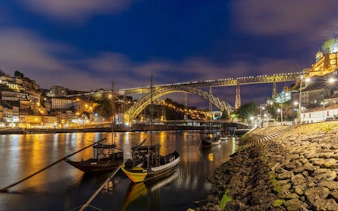 Porto's Dom Luís I Bridge lit up at night over the Douro River with traditional boats.