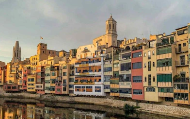 Colorful buildings along the Onyar River in Girona, Spain, with a cathedral in the background.