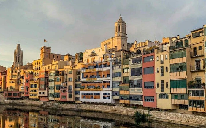 Colorful buildings along the Onyar River in Girona, Spain, with a cathedral in the background.