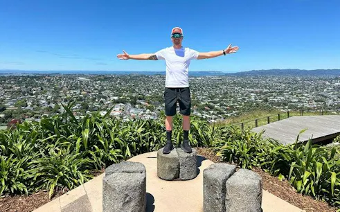 Person standing on stone platform with arms outstretched, overlooking Auckland cityscape.