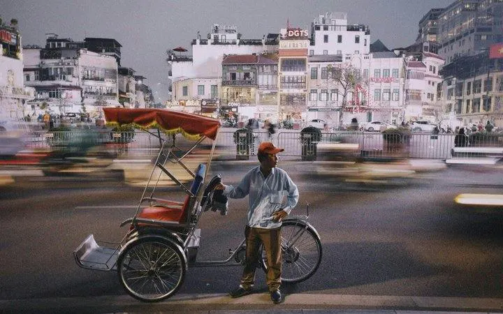Cyclo driver in Hanoi Old Quarter at dusk with blurred city lights.
