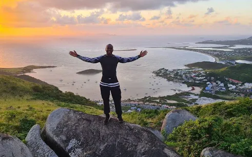 Man standing on a rock with arms outstretched, overlooking St. Maarten's coastline at sunset.