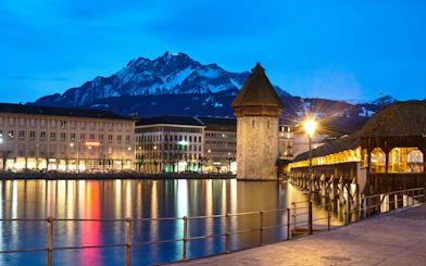 Chapel Bridge and Water Tower at dusk in Lucerne Old Town, with mountains in the background.