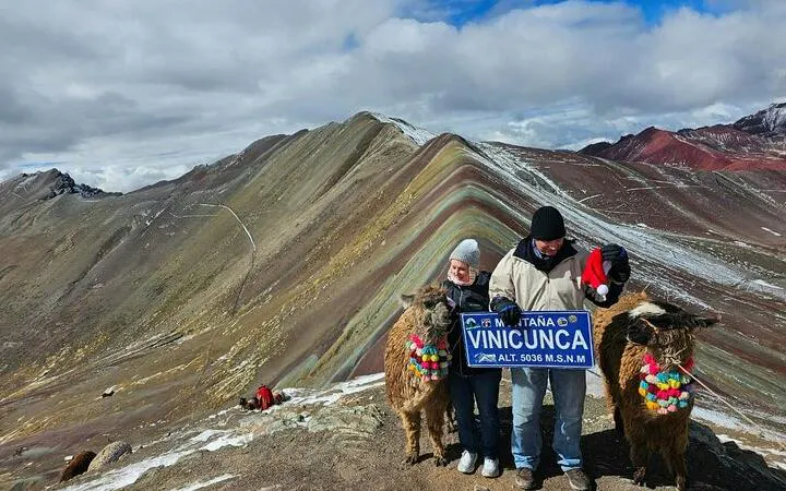 Couple with llamas at Vinicunca Rainbow Mountain, Cusco, holding a sign at 5036m altitude.