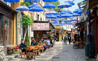 Skopje street with blue umbrellas overhead, cafes, and people walking.
