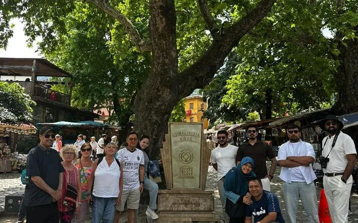 Group of tourists at Cumalıkızık Village in Bursa, standing by a UNESCO sign under a large tree.