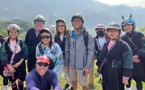 Group of tourists on a Sapa motorbike tour with locals in traditional attire, rice fields in background.