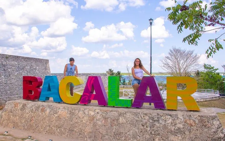 Bacalar sign with visitors, stone wall, and trees under a cloudy sky.