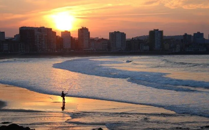 Gijón beach at sunset with a person fishing, city skyline in the background.