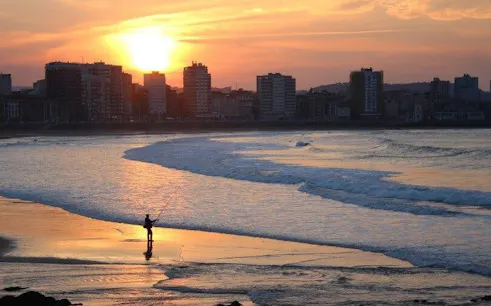 Gijón beach at sunset with a person fishing, city skyline in the background.