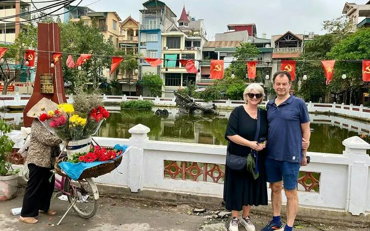 Couple posing by Hanoi lake with flower vendor and red flags in background.