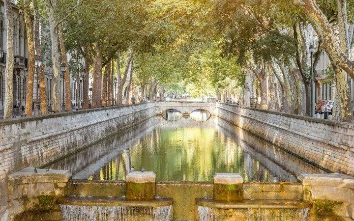 Tree-lined canal in Nîmes with stone bridge, part of self-guided audio walking tour from Arles.