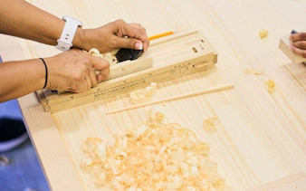 Hands crafting chopsticks in Tokyo Shinjuku workshop with wood shavings and tools.