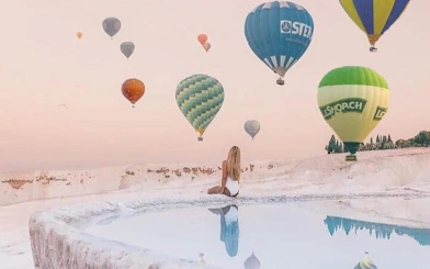 Hot air balloons over Pamukkale travertines with a person sitting by a thermal pool.