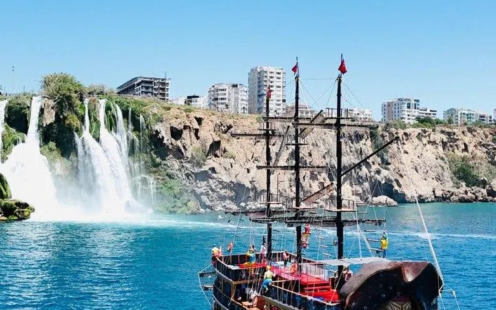 Pirate-themed boat near Lower Duden Waterfall in Antalya with cityscape in background.