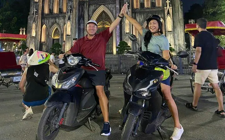 Two people on motorbikes high-fiving in front of St. Joseph's Cathedral, Hanoi at night.