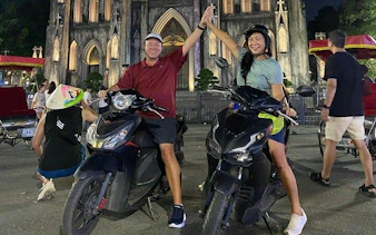 Two people on motorbikes high-fiving in front of St. Joseph's Cathedral, Hanoi at night.