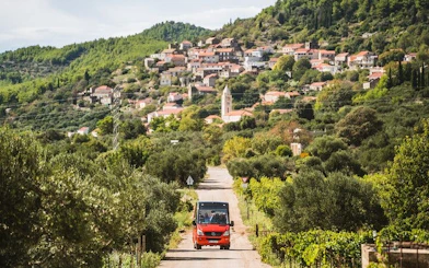 Open-top minibus on a scenic road in Korcula, Croatia, with vineyards and village in the background.