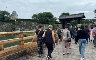 Visitors crossing a wooden bridge towards Himeji Castle, Japan, with stone walls and greenery.