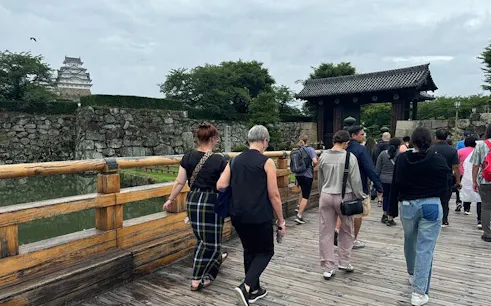 Visitors crossing a wooden bridge towards Himeji Castle, Japan, with stone walls and greenery.