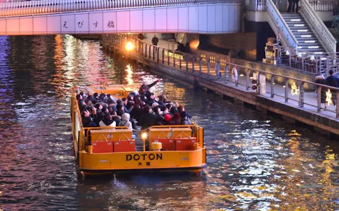 Tourists on a Dotonbori River cruise in Osaka, passing under a lit bridge at night.