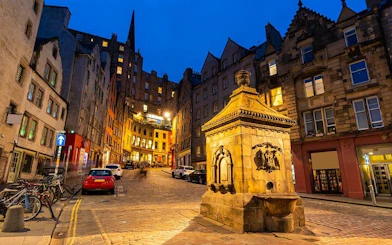 Edinburgh street scene at dusk with historic buildings and a stone monument.