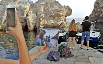 Tourists on Dubrovnik coast with Game of Thrones scene photo, near boats and rocky shore.
