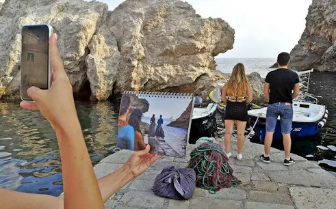 Tourists on Dubrovnik coast with Game of Thrones scene photo, near boats and rocky shore.