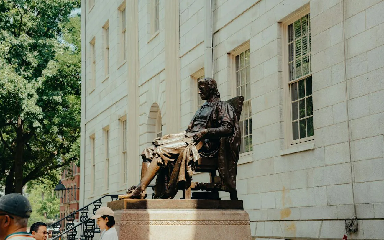 John Harvard Statue in Harvard Yard, Boston, with visitors nearby.