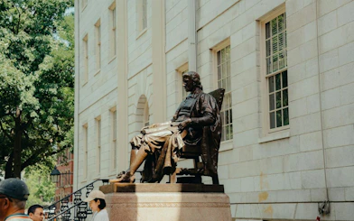 John Harvard Statue in Harvard Yard, Boston, with visitors nearby.