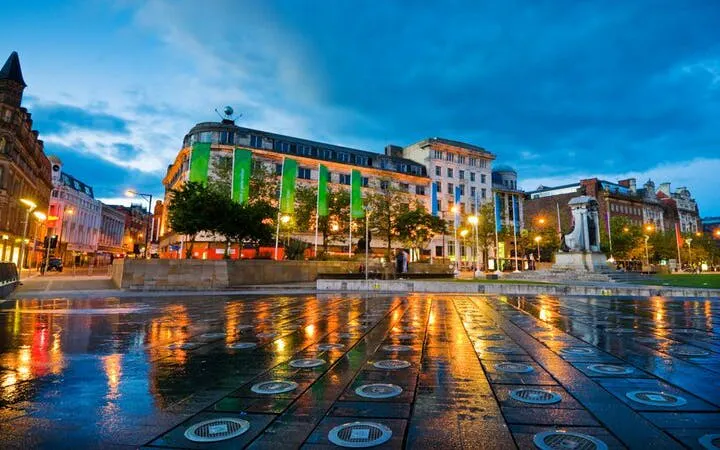 Manchester city square at dusk with illuminated buildings and wet pavement reflecting lights.