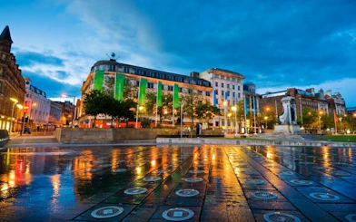 Manchester city square at dusk with illuminated buildings and wet pavement reflecting lights.