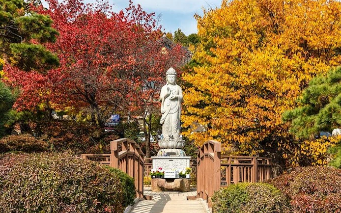 Buddha statue on a wooden bridge surrounded by autumn trees in a Korean garden.