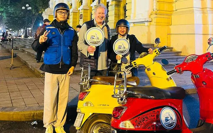 Hanoi Vespa tour participants with scooters in front of a historic building at night.