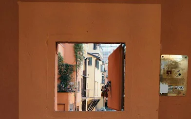 View of Bologna's narrow alley through a small window, showcasing colorful buildings.