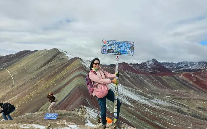 Person standing by a sign at Rainbow Mountain, with colorful peaks in the background, Cusco.