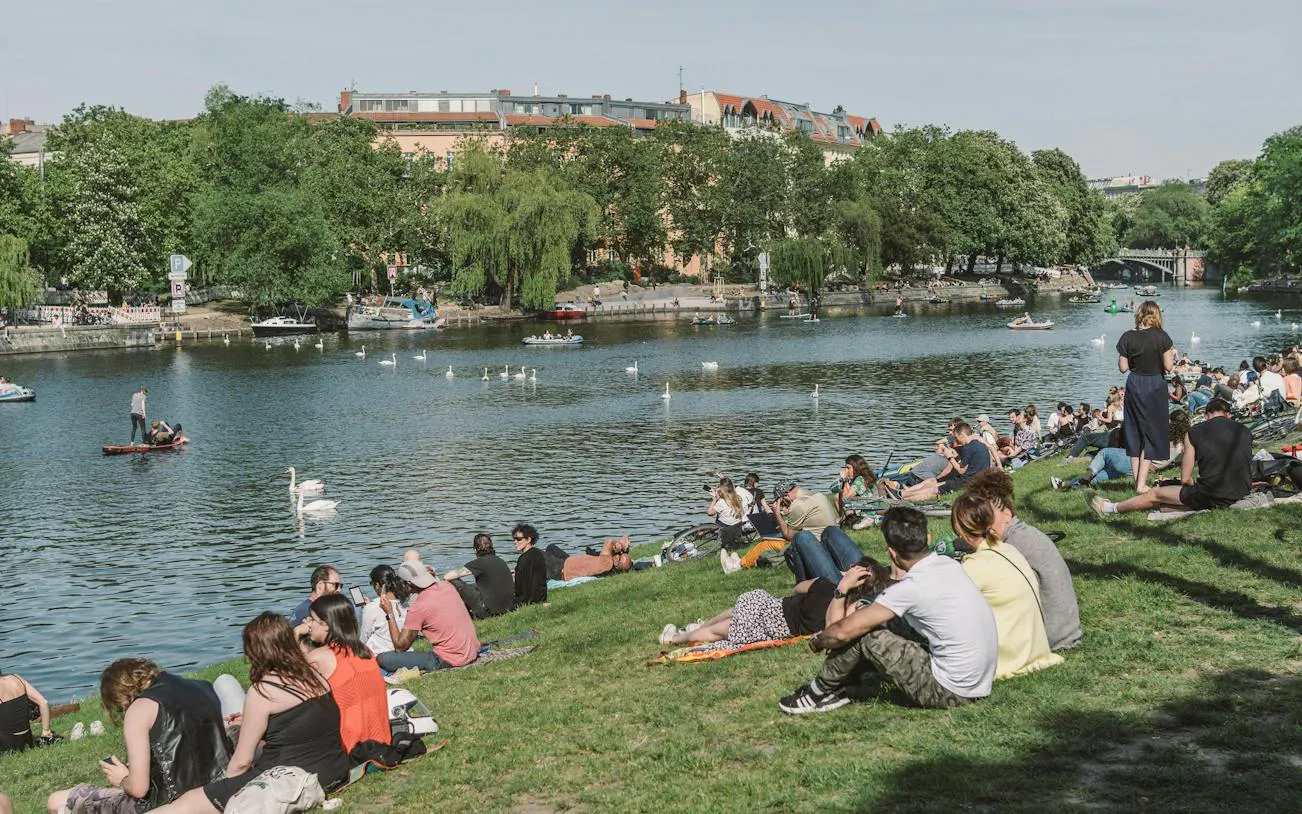 People relaxing by the Landwehr Canal in Kreuzberg, Berlin, with swans and boats on the water.
