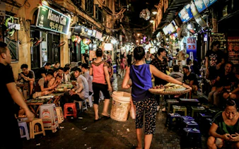 Hanoi Old Quarter night market with people dining and a vendor carrying fruit.