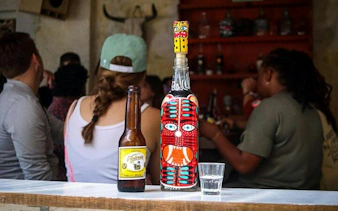 Bottle with colorful mask design and beer on bar counter, Mexico City taco tour.