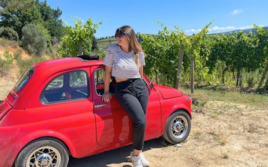 Vintage red Fiat 500 parked by vineyards in Chianti, Italy, with a person sitting on the car.
