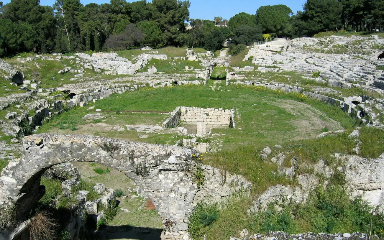 Roman Amphitheatre ruins at Neapolis Archaeological Park, Siracusa, surrounded by greenery.