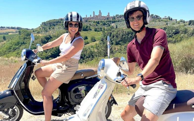 Couple riding Vespas through Chianti countryside with San Gimignano in the background.