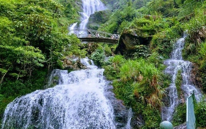 Waterfall with bridge in lush greenery, part of Sapa motorbike tour to Pusamcap Cave.