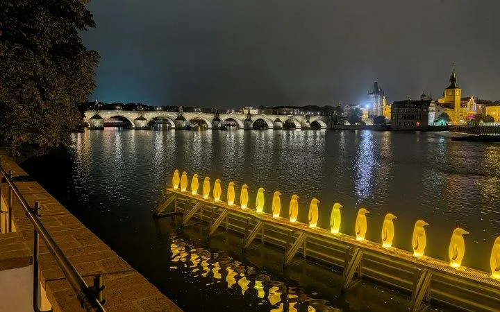 Prague night view with illuminated penguin sculptures on Vltava River, Charles Bridge in background.