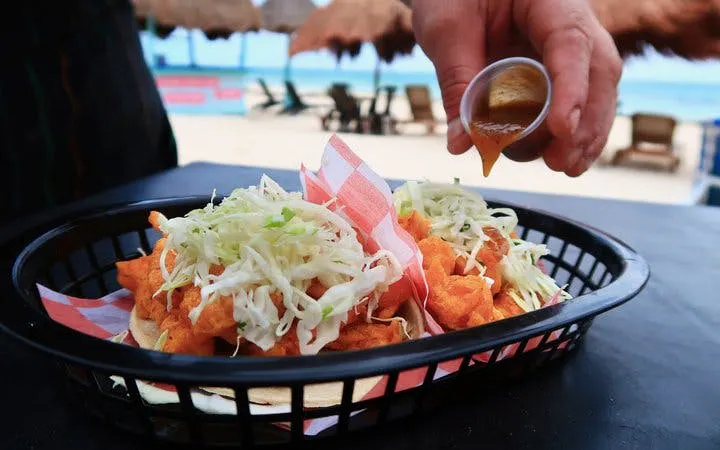 Tacos with cabbage slaw being garnished with sauce on a beachside table in Mexico.