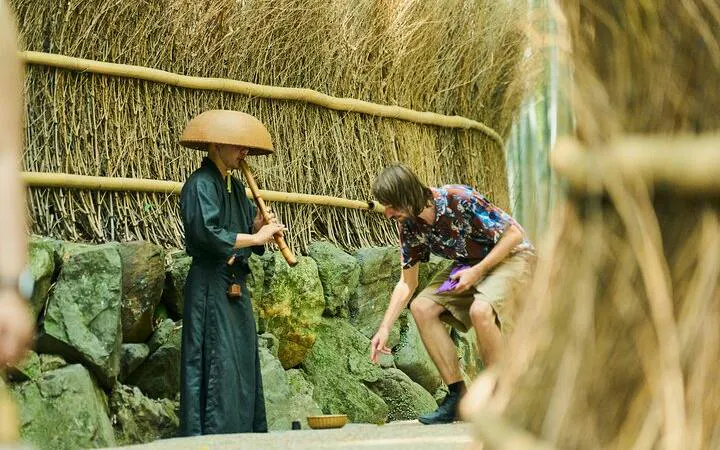Musician playing flute in Arashiyama bamboo grove, Kyoto, with tourist observing.