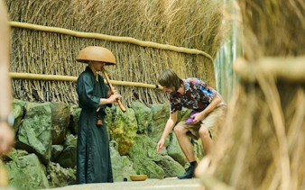 Musician playing flute in Arashiyama bamboo grove, Kyoto, with tourist observing.